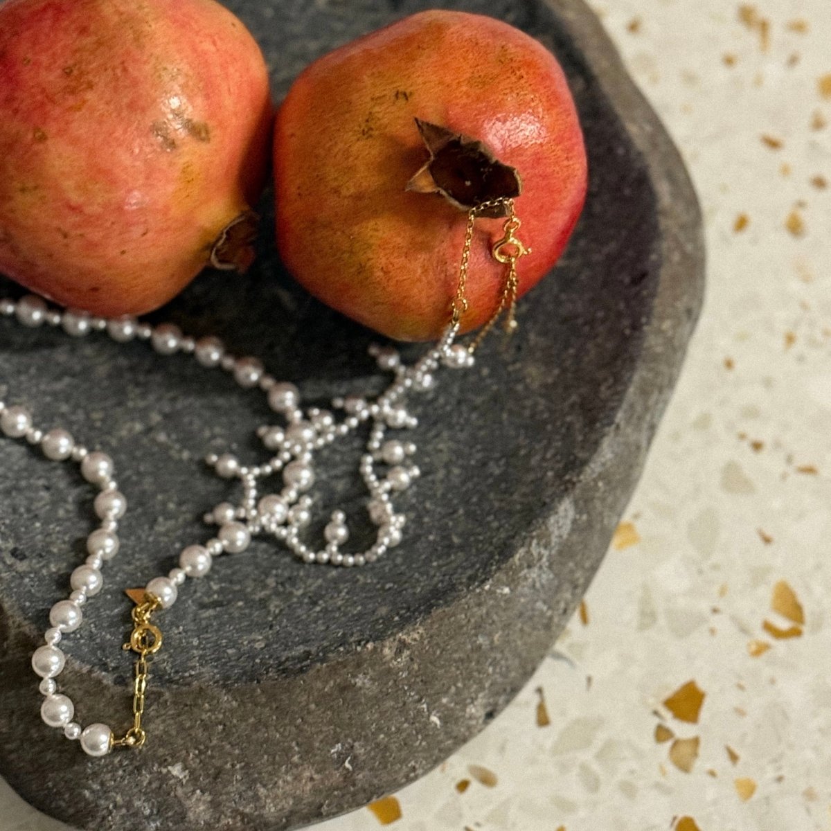 Aegean Pearl choker and Sacred Pearl necklace in a stone basin beside of two pomegranates