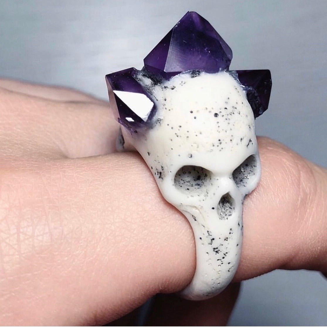 Bifacial Skull Ring on a model's hand, showing one of the skulls and rich-purple, gleaming amethyst crystals, close up