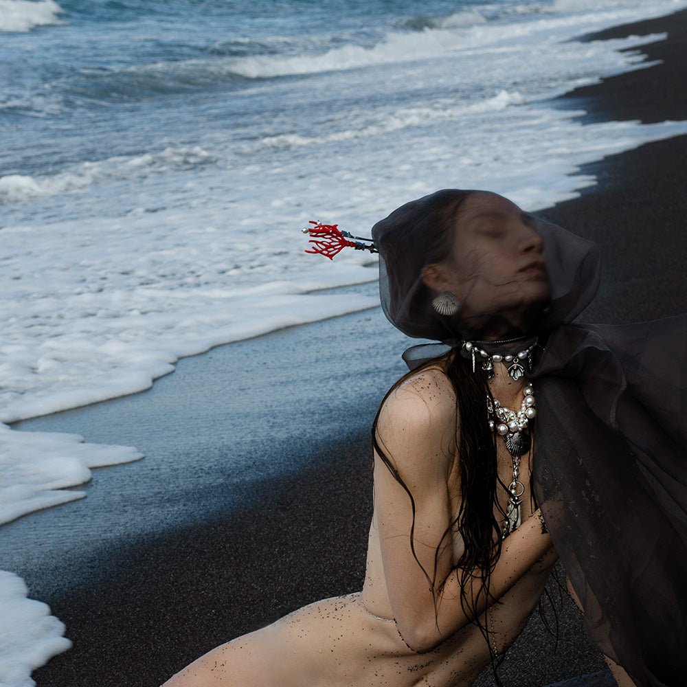 Siren Pearl Necklace - fashion editorial, female model on a sea shore with black sand and foamy waves wearing several pieces including siren pearl necklace