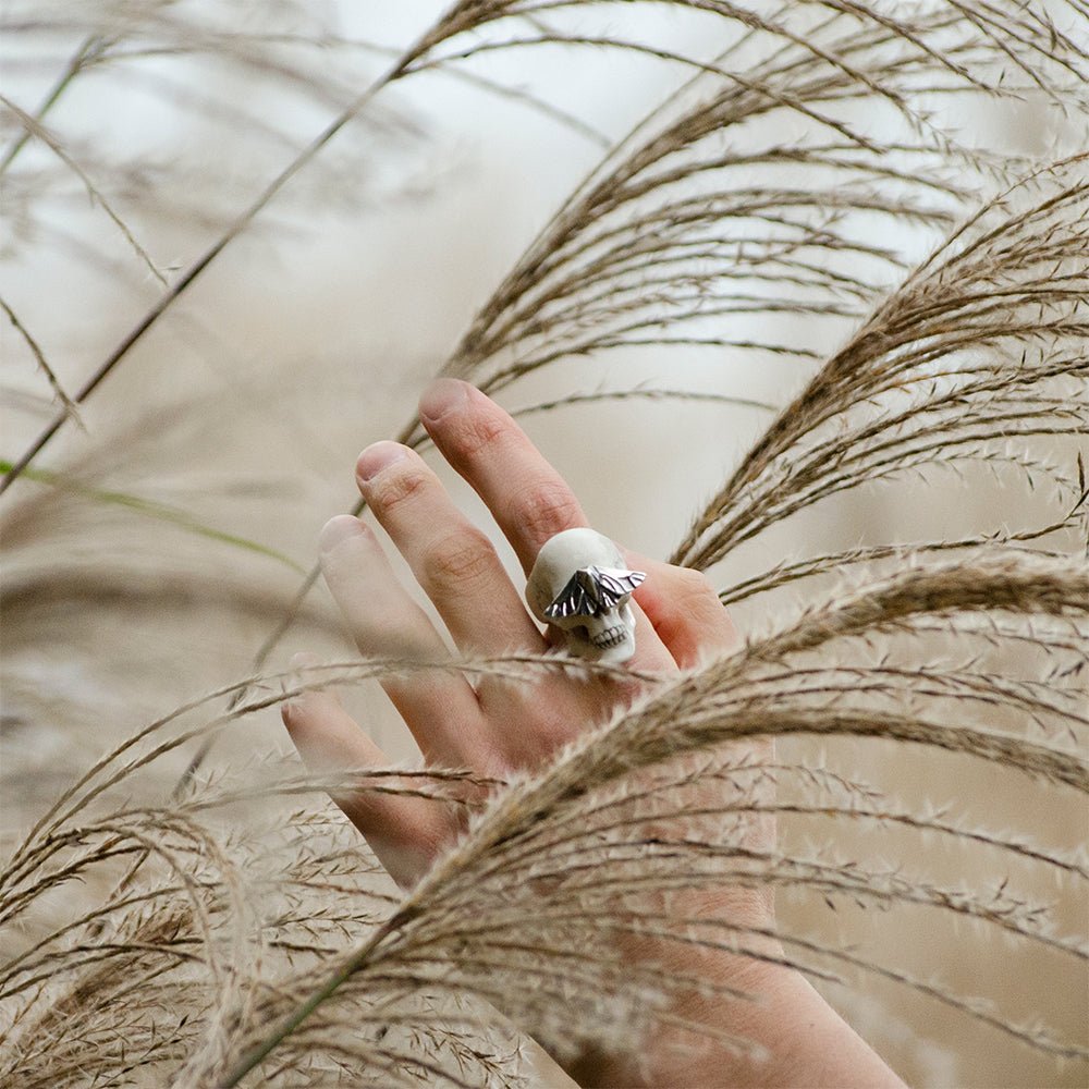 Skull & Moth ring - white variant - on a model's hand reaching through tall autumnal grass, pastel palette, image has melancholic mood